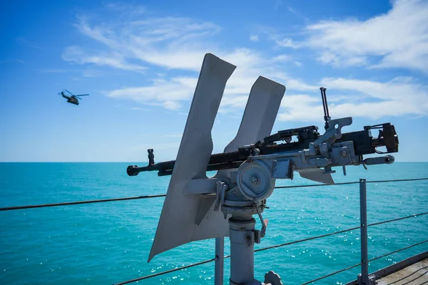 Color image of an automated machine gun on the deck of a military ship, at sea, and a flying helicopter.