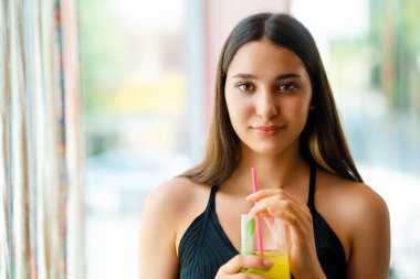 Pencerenin önünde gülümseyen ve içecek içen genç bir kadın..