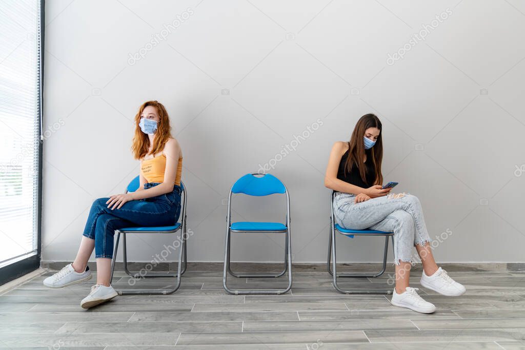 Two young woman with protection mask is sitting in a waiting hall with a distance. Corona virus outbreaking.