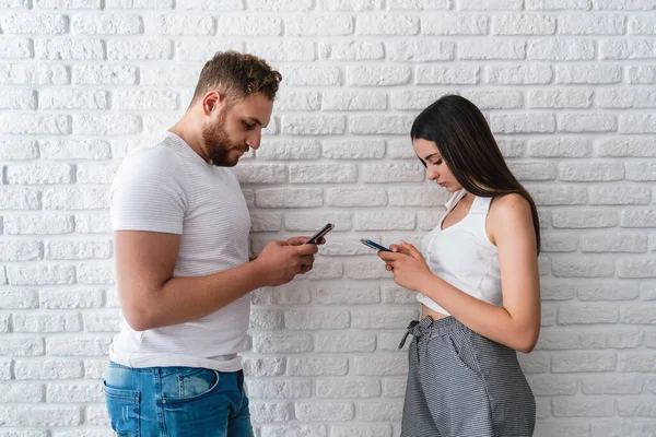 Two friends or lovers standing front of awall and looking to their mobile phones. Technology addiction concept. — Stock Photo, Image