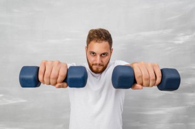 Beyaz tişörtlü genç adam gri duvara karşı halterli egzersiz yapıyor.