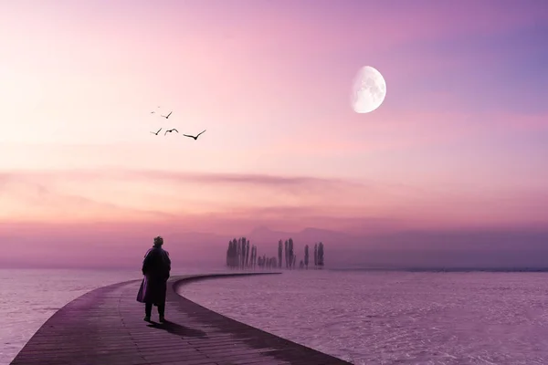 Un homme solitaire se tient sur une jetée en bois et regarde vers l'horizon avec un ciel lilas et la lune — Photo