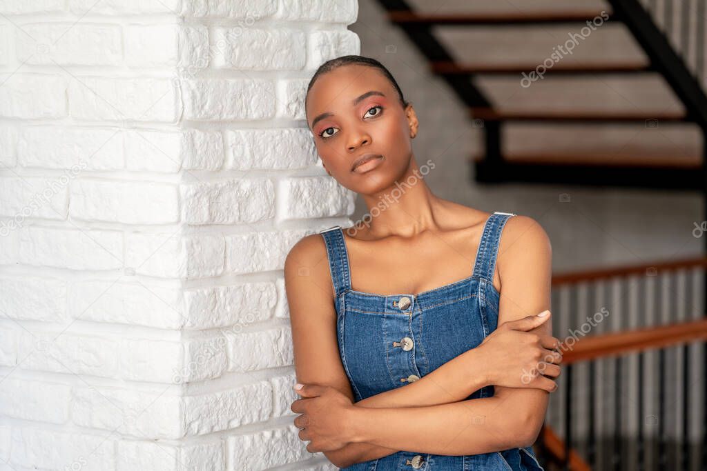 Young dark skin woman leans back on a white brick wall with crossed arms and looking to a point.