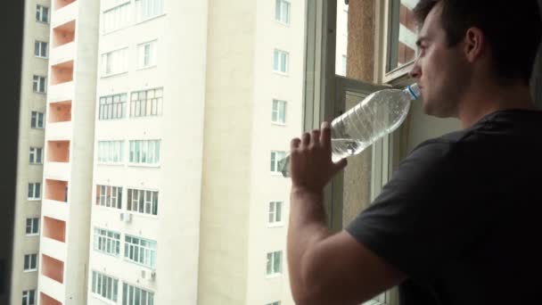 Άνθρωπος πόσιμο νερό από ένα μπουκάλι στέκεται δίπλα στο παράθυρο — Αρχείο Βίντεο
