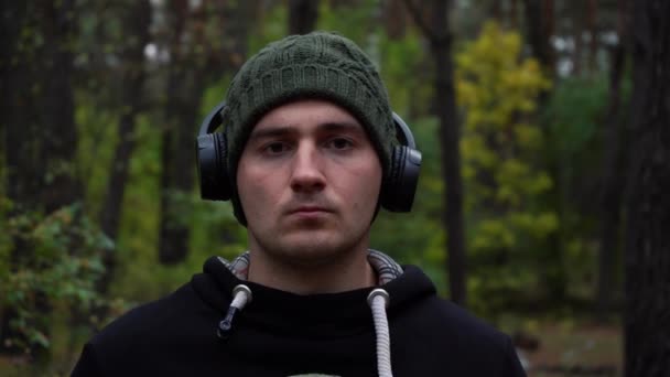 Portret van een mannelijke atleet kijkend naar de camera in het midden van het bos in een hoed en een koptelefoon — Stockvideo