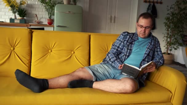 Seorang pria berkacamata membaca sebuah buku tergeletak di sofa Moscow Russia sekote 20, 2021 — Stok Video