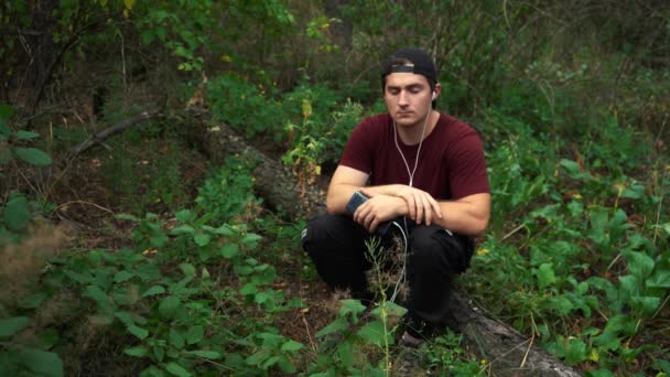 Um homem ouve música enquanto está sentado em uma árvore derrubada na floresta — Vídeo de Stock