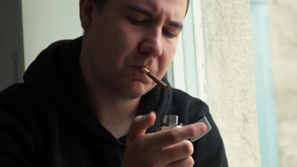 En man tänder en cigarett när han sitter på fönsterbrädan i sitt rum. — Stockvideo