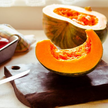 Parçalara ayrılmış parlak turuncu bir balkabağı fırın kabında ve bıçak ve kırmızı elmaların yanında doğrama tahtasında yatıyor.