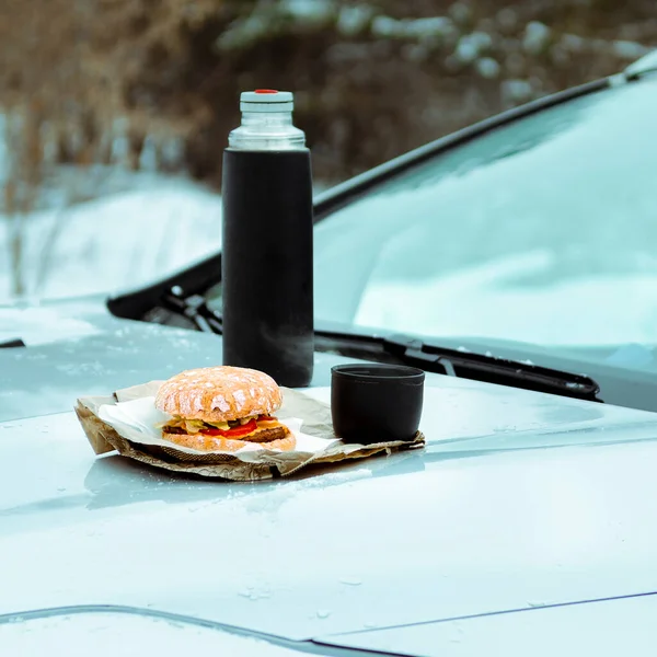 Winter picnic with tea and burger on the hood of a silver car against the background of the winter forest