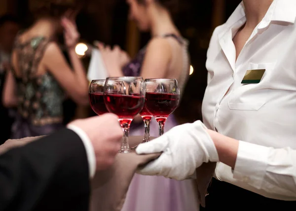 Close-up picture of waiter's hands wearing white gloves holding a tray with red wine, serving alcohol drinks. Catering service at special occasion, event. Hospitality industry concept.