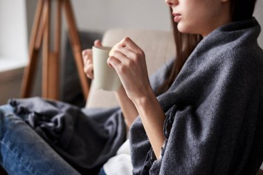 27 Feb 2019 Ternopil, Ukraine. Close-up picture of young woman, holding mug of coffee tea, sitting in armchair, covered with blanket. Snug atmosphere. Sweet food consumption. Day off relaxation
