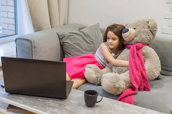 Child girl watching laptop screen, hugging teddy bear. Concept of distance learning, e-learning, and leisure of modern children at computer.