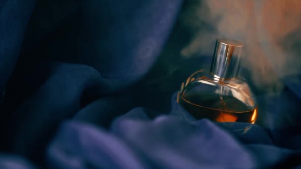 Botella de perfume sobre tela azul en nube de humo. Primer plano de vídeo en cámara lenta. — Vídeos de Stock