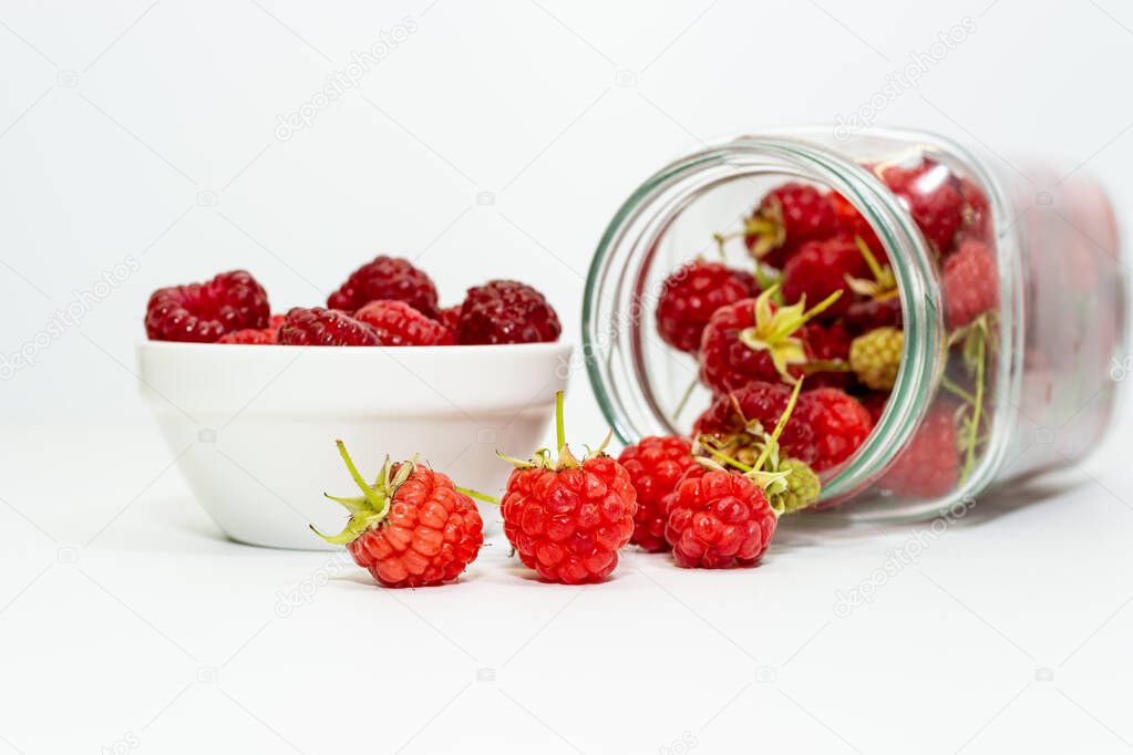 Freshly picked red raspberries in a small bowl and in an inverted glass jar on a white background.