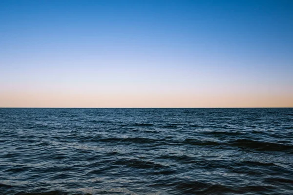 Dawn over the horizon of the dark waters of the ocean. Small waves and a blue clear sky without clouds against the background of a yellow-orange glow.