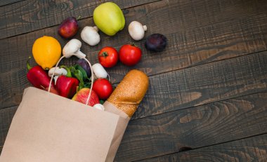 Koyu ahşap arka planda çeşitli taze sebzeler ve ekmekle dolu bir kağıt alışveriş torbası. Alışveriş konsepti. Sağlıklı organik seçimler. Üst görünüm, boşluğu kopyala. Alışveriş için telefon ve kredi kartı..