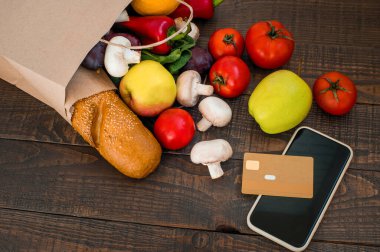 Koyu ahşap arka planda çeşitli taze sebzeler ve ekmekle dolu bir kağıt alışveriş torbası. Alışveriş konsepti. Sağlıklı organik seçimler. Üst görünüm, boşluğu kopyala. Alışveriş için telefon ve kredi kartı..