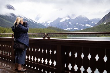 Yetişkin sarışın kadın Buzul Ulusal Parkı Montana ABD 'deki Many Buzul Oteli' nin güvertesinde Swiftcurrent Gölü 'nün fotoğraflarını çekiyor.