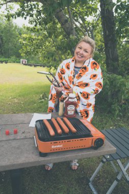 Montana, USA - July 4, 2022: Woman grills hot dogs on an outdoor camp stove using tongs, at a picnic site, wearing a pumpkin blanket