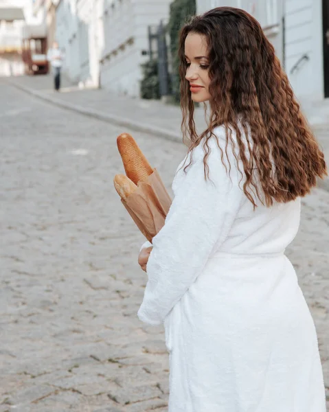 白い衣を着た暗い髪の若い女性が街の通りを見ている 若いスタイリッシュな女性がワルシャワ市内の通りに立っているフランスのバゲットを購入 — ストック写真