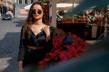 Şehir caddesinde çiçek çalılarının yanında poz veren güneş gözlüklü şirin bir model. Uzun saçlı, siyah elbiseli, güzel bir kadın, eski şehir sokaklarında çiçek açan çalıların yanında poz veriyor..