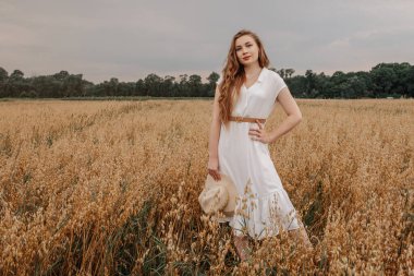 Beyaz elbiseli ve şapkalı güzel bir kız tarlada yürüyor ve günbatımında yulaf pozu veriyor. Tarlada mutlu bir genç kadın portresi. Yulaf tarlası.