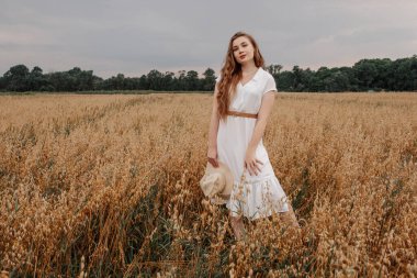 Beyaz elbiseli ve şapkalı güzel bir kız tarlada yürüyor ve günbatımında yulaf pozu veriyor. Tarlada mutlu bir genç kadın portresi. Yulaf tarlası.