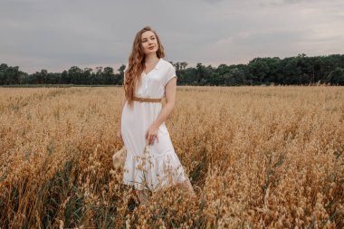 Beyaz elbiseli ve şapkalı güzel bir kız tarlada yürüyor ve günbatımında yulaf pozu veriyor. Tarlada mutlu bir genç kadın portresi. Yulaf tarlası.