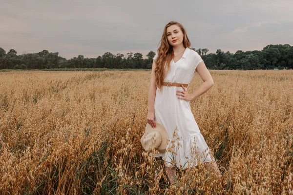Beyaz elbiseli ve şapkalı güzel bir kız tarlada yürüyor ve günbatımında yulaf pozu veriyor. Tarlada mutlu bir genç kadın portresi. Yulaf tarlası.