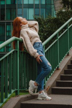 Sokak merdiveninde kızıl saçlı kadın. Şık giyinmiş kızıl saçlı kadın. Uzun kızıl saçlı, modacı kız. Sokak stili, modern, katı şehir kızı merdivenlerde. Şehirde moda yürüyüşü..