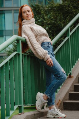 Sokak merdiveninde kızıl saçlı kadın. Şık giyinmiş kızıl saçlı kadın. Uzun kızıl saçlı, modacı kız. Sokak stili, modern, katı şehir kızı merdivenlerde. Şehirde moda yürüyüşü..