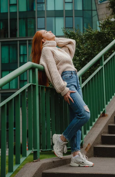 Sokak merdiveninde kızıl saçlı kadın. Şık giyinmiş kızıl saçlı kadın. Uzun kızıl saçlı, modacı kız. Sokak stili, modern, katı şehir kızı merdivenlerde. Şehirde moda yürüyüşü..
