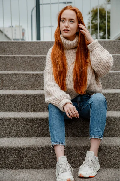 Günbatımı portresindeki genç kadın güneş ışığıyla. Kızıl saçlı, kot pantolon giyen, merdivenlerde oturan güzel, genç ve şık bir kadın. Sokakta ateş eden mutlu genç bayan. Polonya.