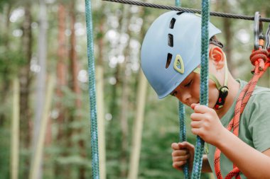 Cesur küçük çocuk macera parkında eğleniyor ve kaskla kameraya gülümsüyor. İp atma alıştırması yapan bir izci. Çocuklar. İp parkında bir kız ve bir erkek engelleri aşıyor. Seyahat.