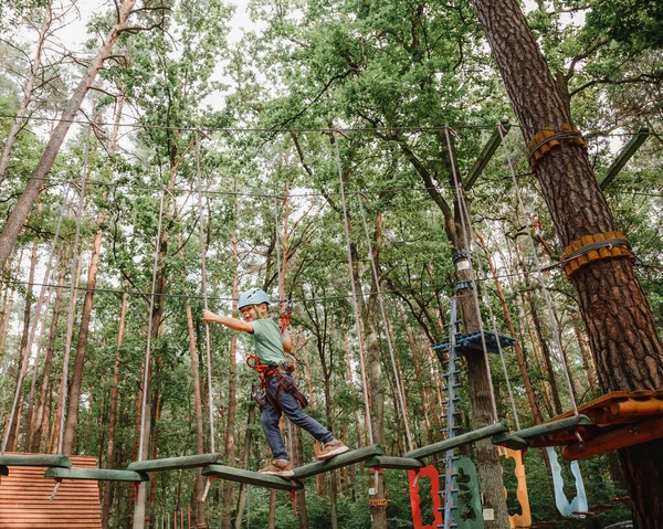 Brave little boy having fun at adventure park and smiling to camera wearing helmet. Scout practicing rappelling. Child - a boy and a girl in the rope park pass obstacles. — Fotografia de Stock