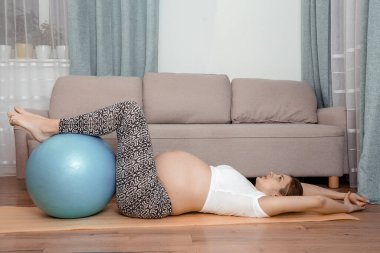 Güzel hamile bir kadın aerobik topuyla jimnastik salonunda. Yoga minderinde hamile bir kadın. Evdeki saydam fitness topunda bacakları tutuyor..