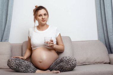 Odadaki kanepede bir bardak suyla oturan hamile bir kadın. Su içen güzel hamile bir kadın. Hamile kadının fotoğrafı..
