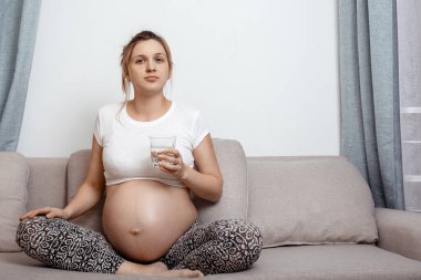 Odadaki kanepede bir bardak suyla oturan hamile bir kadın. Su içen güzel hamile bir kadın. Hamile kadının fotoğrafı..