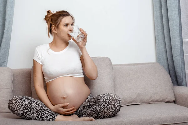 Pregnant woman with a glass of water sitting on sofa in the room. Pretty pregnant woman drinking water. Photo of pregnant woman.
