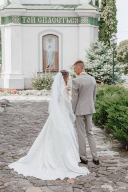Happy wedding photography of bride and groom at wedding ceremony. The groom looks at the bride. Bride and groom holding hands, walk near the church after the wedding ceremony. Wedding day.