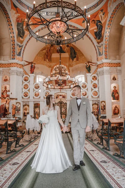 Happy wedding photography of bride and groom at wedding ceremony. The groom looks at the bride. Bride and groom holding hands, walk near the church after the wedding ceremony. Wedding day.