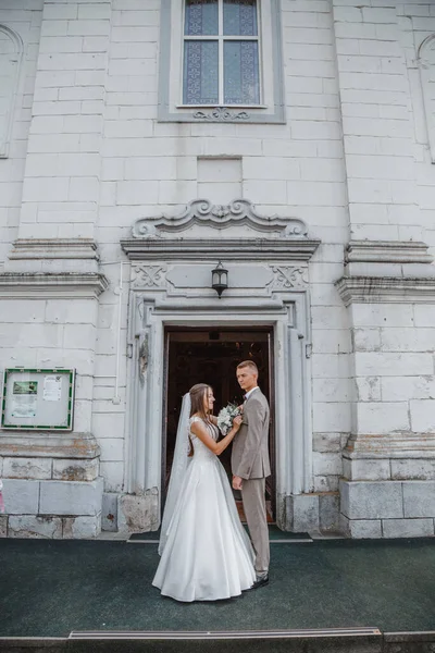 Happy wedding photography of bride and groom at wedding ceremony. The groom looks at the bride. Bride and groom holding hands, walk near the church after the wedding ceremony. Wedding day.