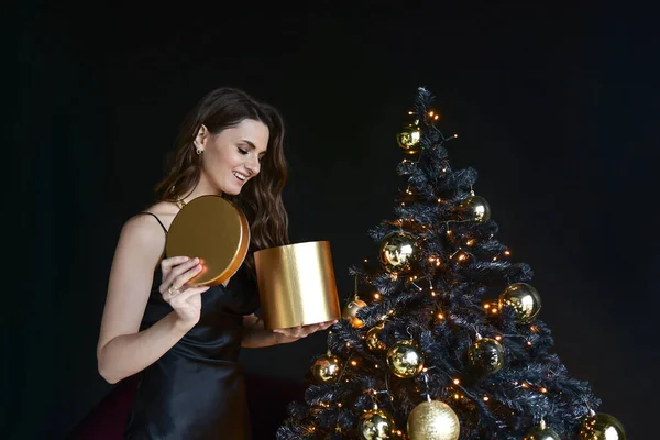 クリスマスツリーを背景に美しい若い女性。新年の贈り物だ。黒の背景. — ストック写真