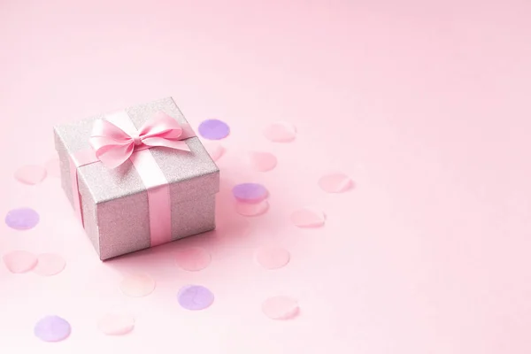 Zilveren glitter geschenkdoos met roze lint strik op roze achtergrond met confetti. — Stockfoto