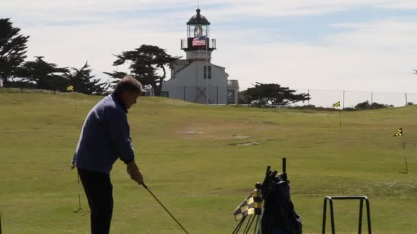 Mann schlägt Golfball mit Leuchtturm eine amerikanische Flagge in der Ferne — Stockvideo