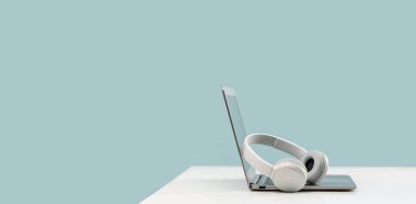 Dizüstü bilgisayar. Beyaz masadaki kablosuz kulaklıklar ve model ekran. Düz mavi arka plan. Uzaktan öğrenme. Evden çalışmak, online kurslar. Çok az. Yardım masası desteği veya çağrı merkezi kulaklığı