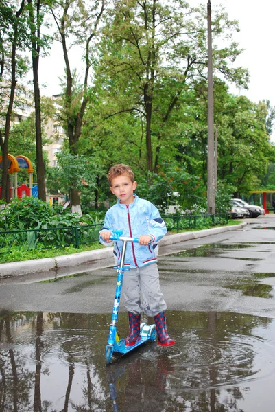 Um menino montando uma scooter através de poças . — Fotografia de Stock