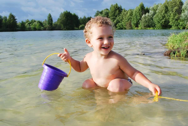 Menino sentado no lago, e brinca com um balde e um ancinho . — Fotografia de Stock