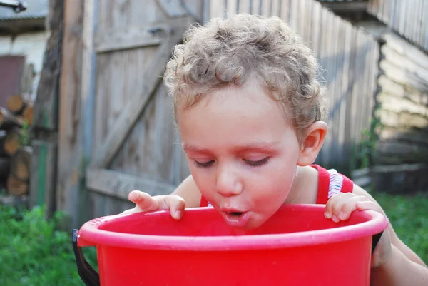 Im Dorf in der Nähe des Brunnens trinkt ein kleiner Junge Wasser aus einem — Stockfoto